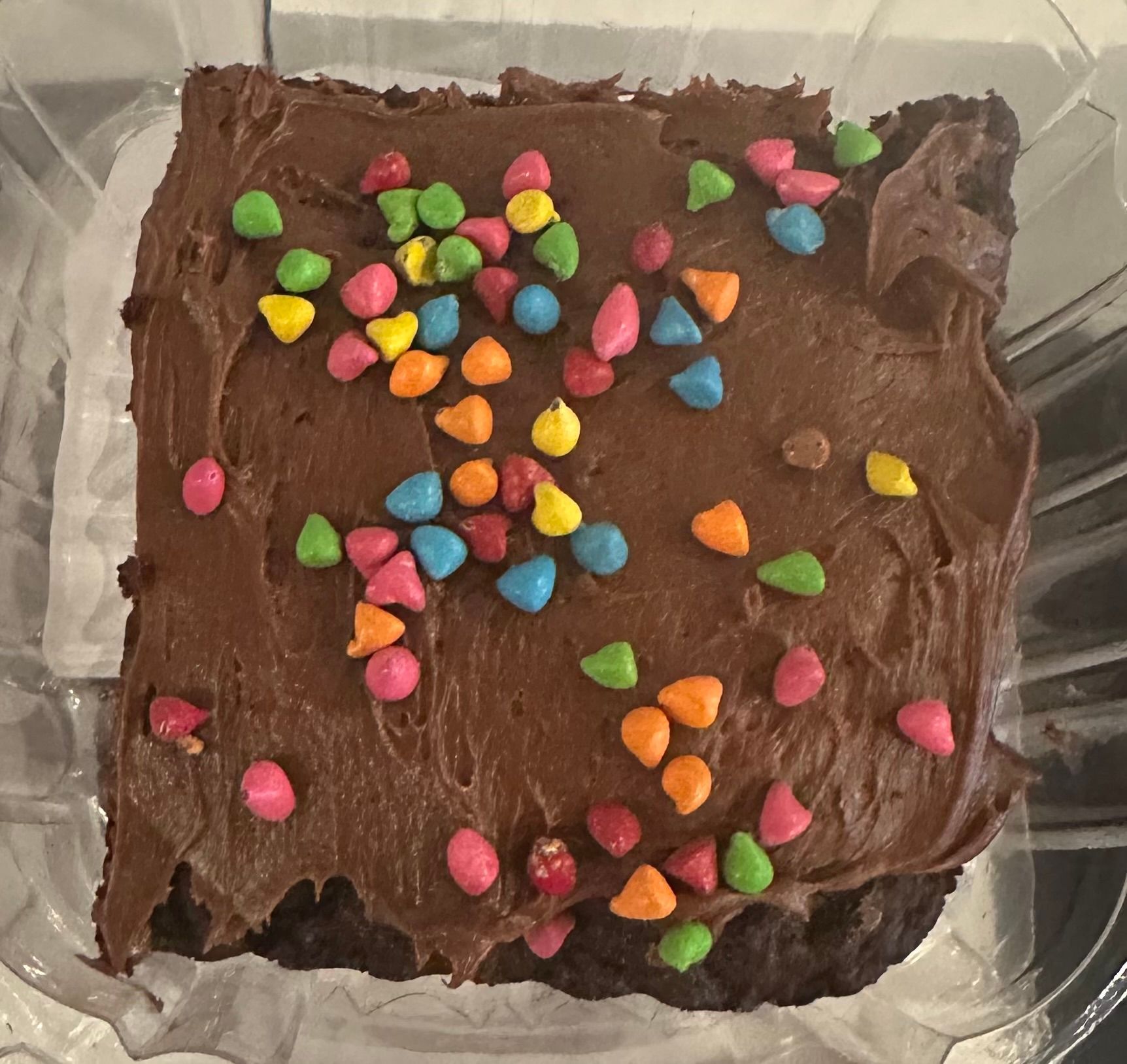 Chocolate frosted cake in a clear container, topped with colorful candy sprinkles