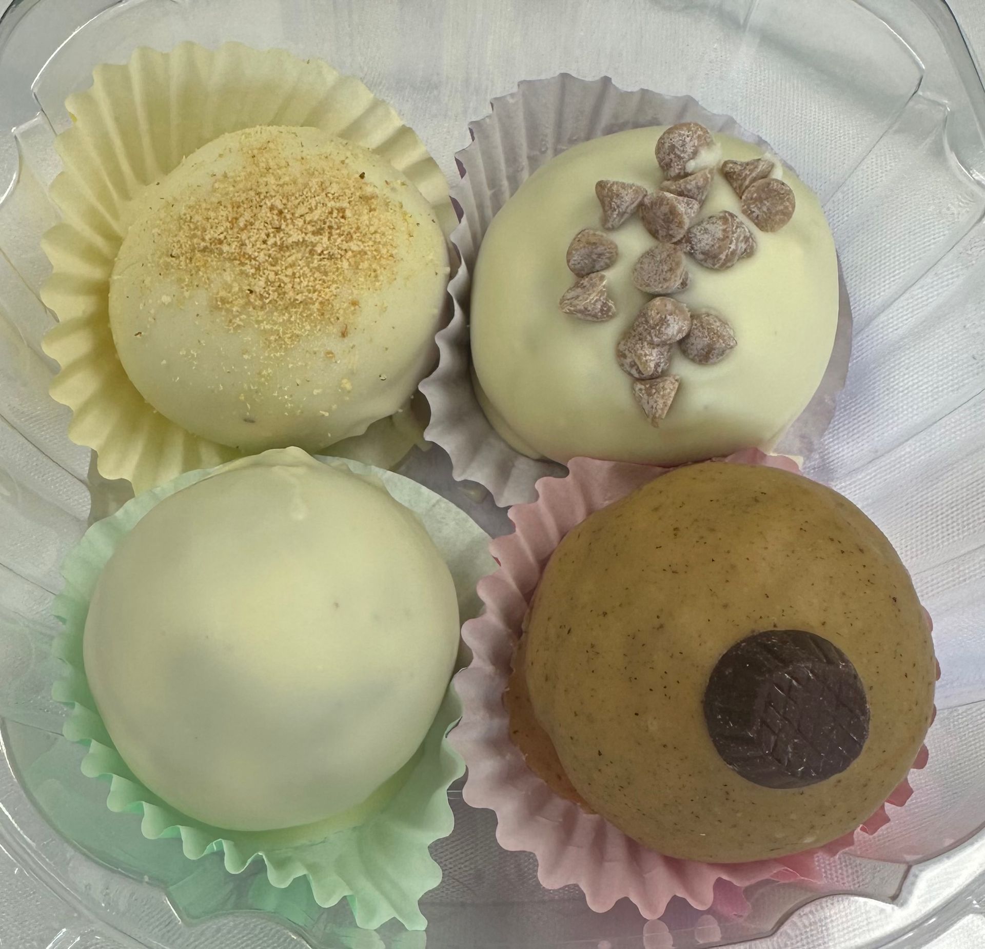 Four assorted round desserts in pastel paper cups, including white-glazed and brown-topped treats.