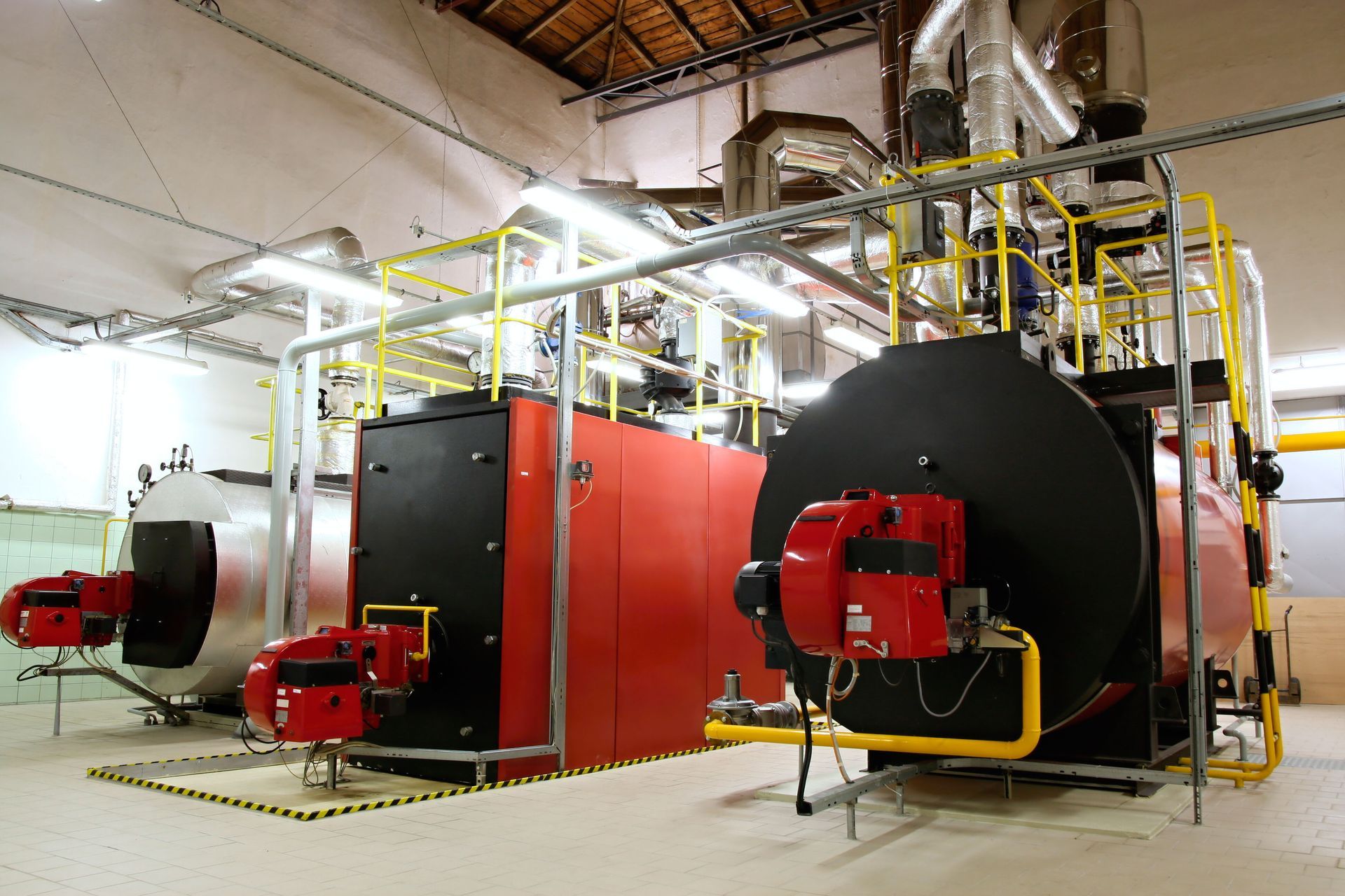 Three industrial boilers in a room, two red and one silver. Pipes and yellow railings are visible.