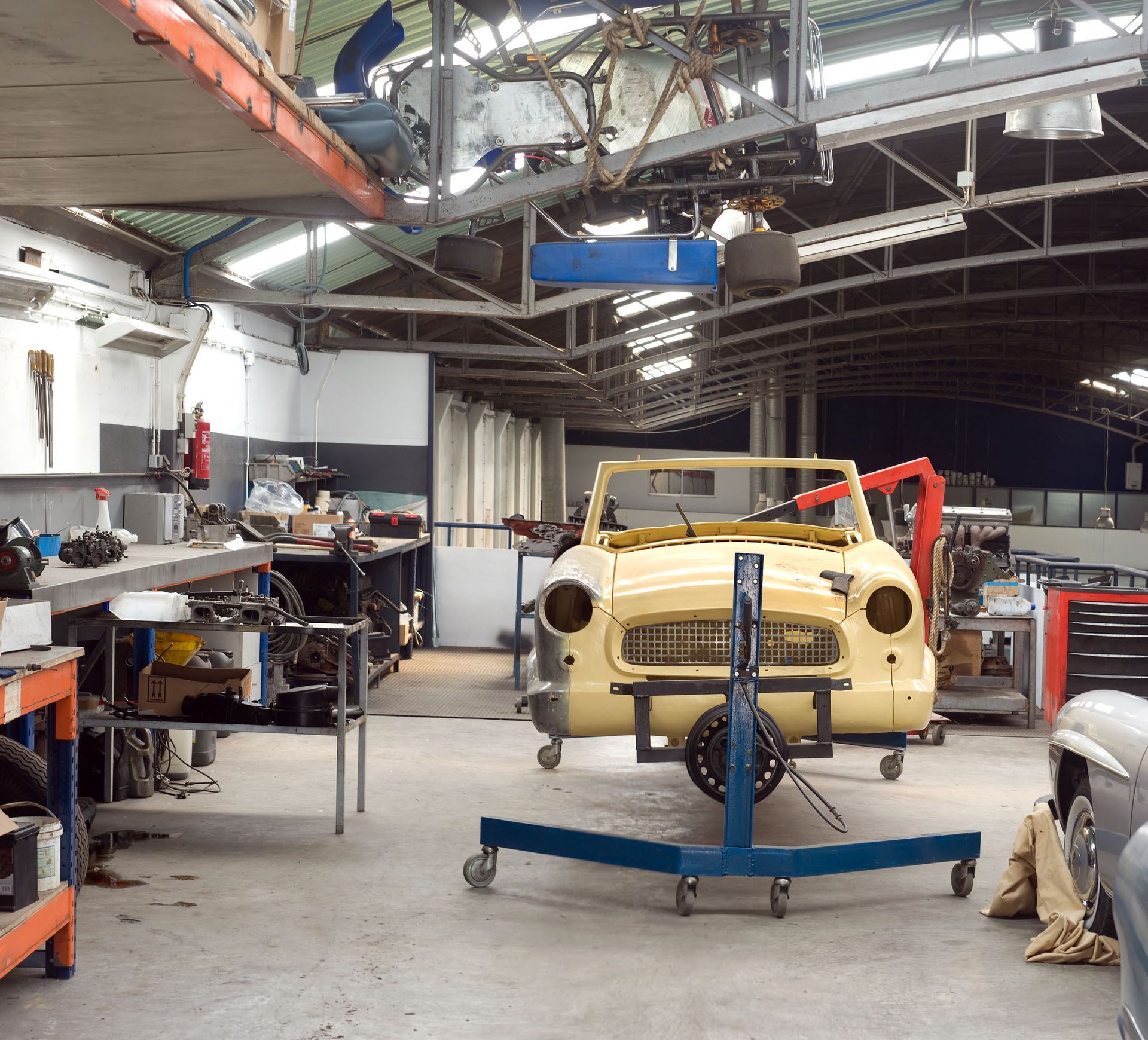 Car repair workshop interior, classic yellow car chassis on a wheeled lift. Tools, parts, and other vehicles visible.