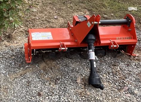 Orange rotary tiller on gravel, ready to attach to a tractor.