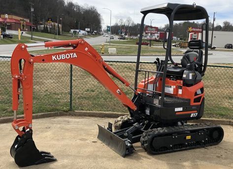 A small orange and black kubota excavator is parked in front of a chain link fence.