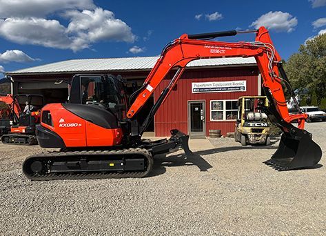 Orange Kubota excavator parked in front of a red building with a sign. A forklift is to the right.