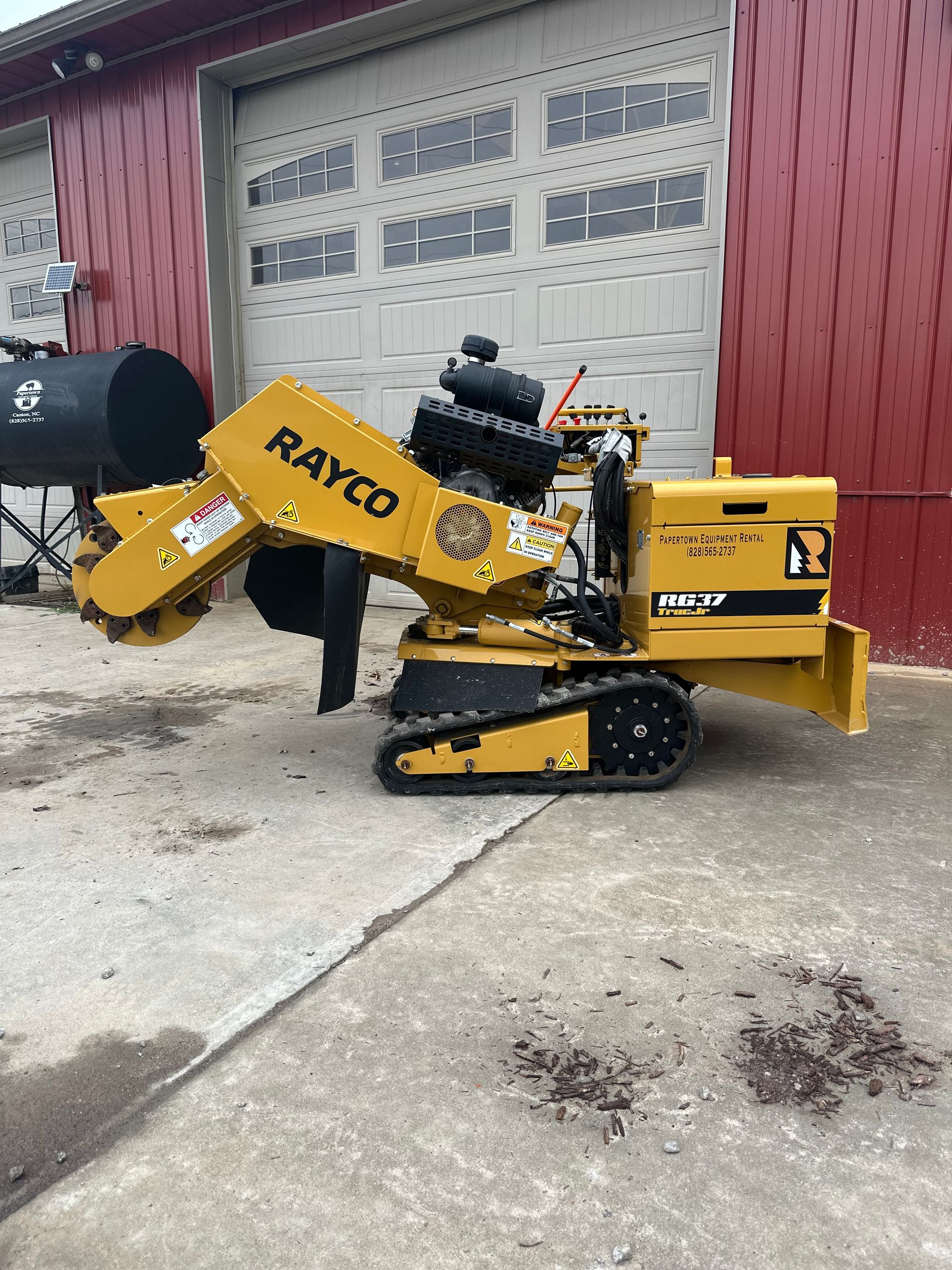 Yellow Rayco stump grinder on tracks, parked in front of a red building with a roll-up door.