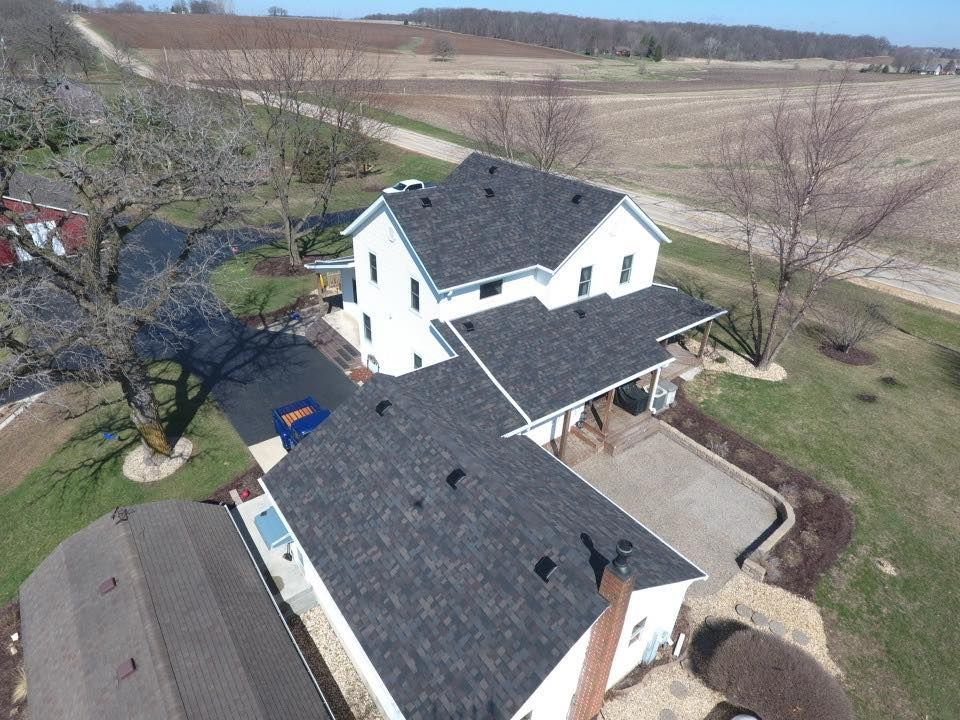 Aerial view of a white house with a dark gray roof, driveway, and surrounding farmland.
