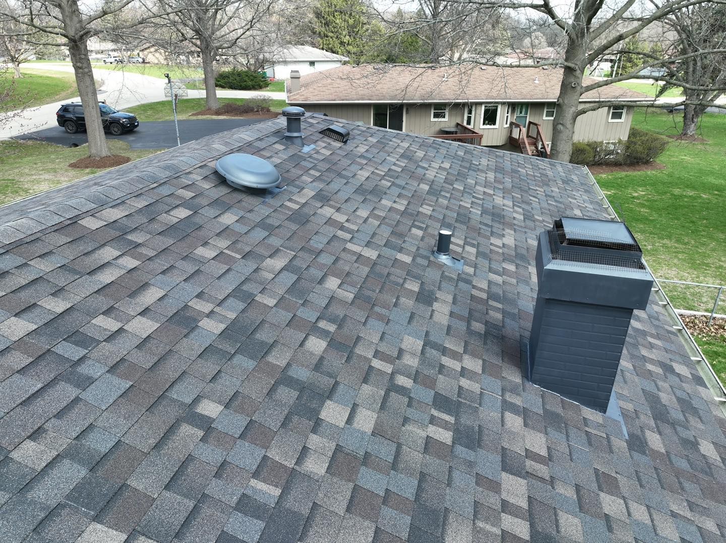 Roof with gray and brown shingles, vents, and chimney in a residential neighborhood.