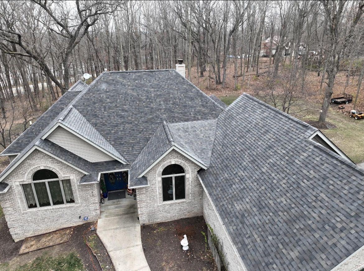 Gray brick house with dark gray shingled roof, trees in background.