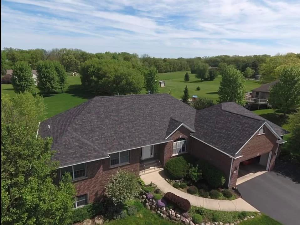 Aerial view of a brick house with a dark gray roof, driveway, and lush green yard surrounded by trees under a blue sky.