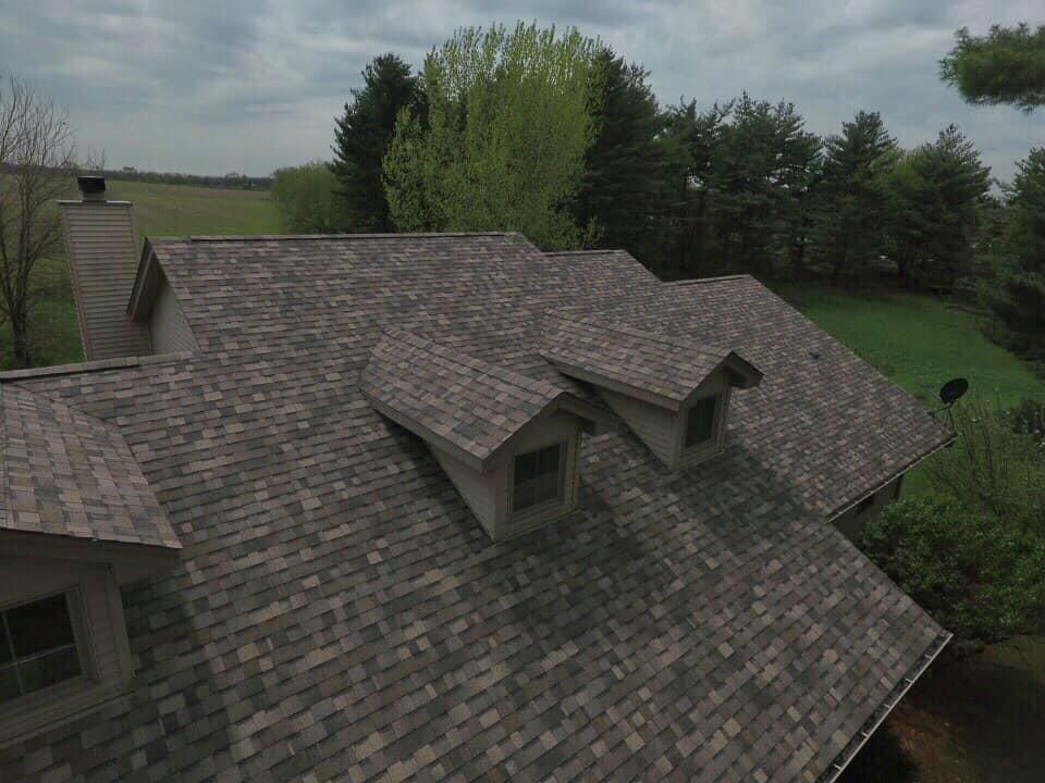 Overhead view of a house with a brown asphalt shingle roof, dormers, and chimney, surrounded by greenery.