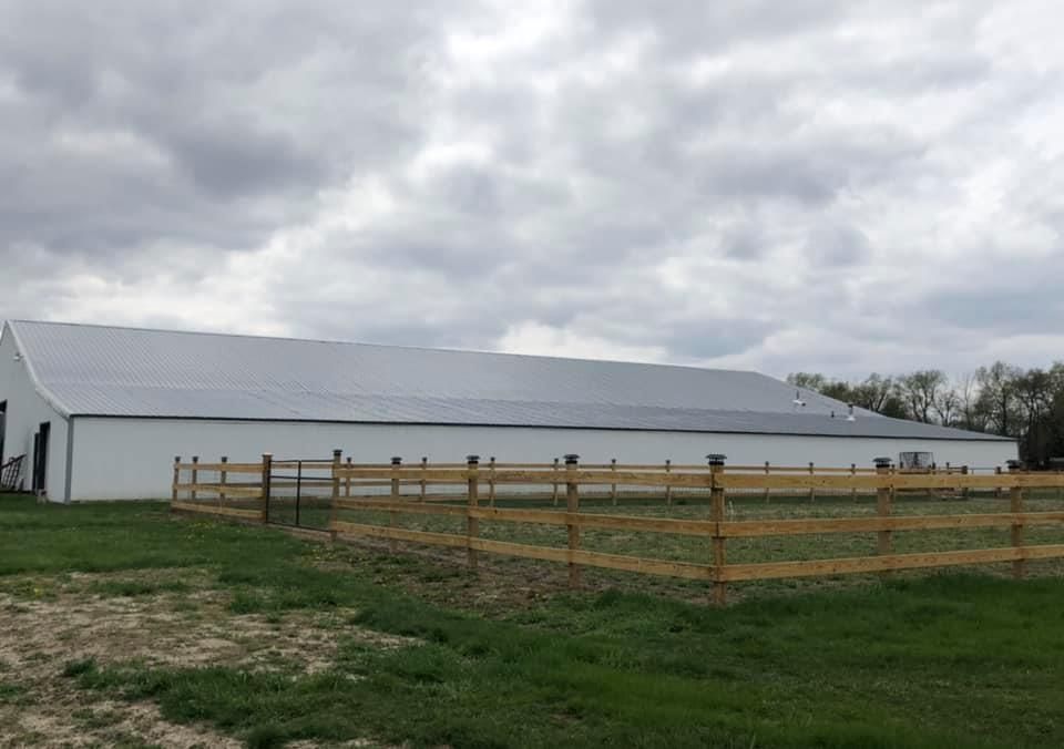 White barn with gray roof covered in solar panels, wooden fence, and green grass under a cloudy sky.