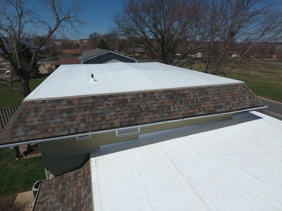 Aerial view of a house with a flat white roof and brown shingled side with a clear sky.