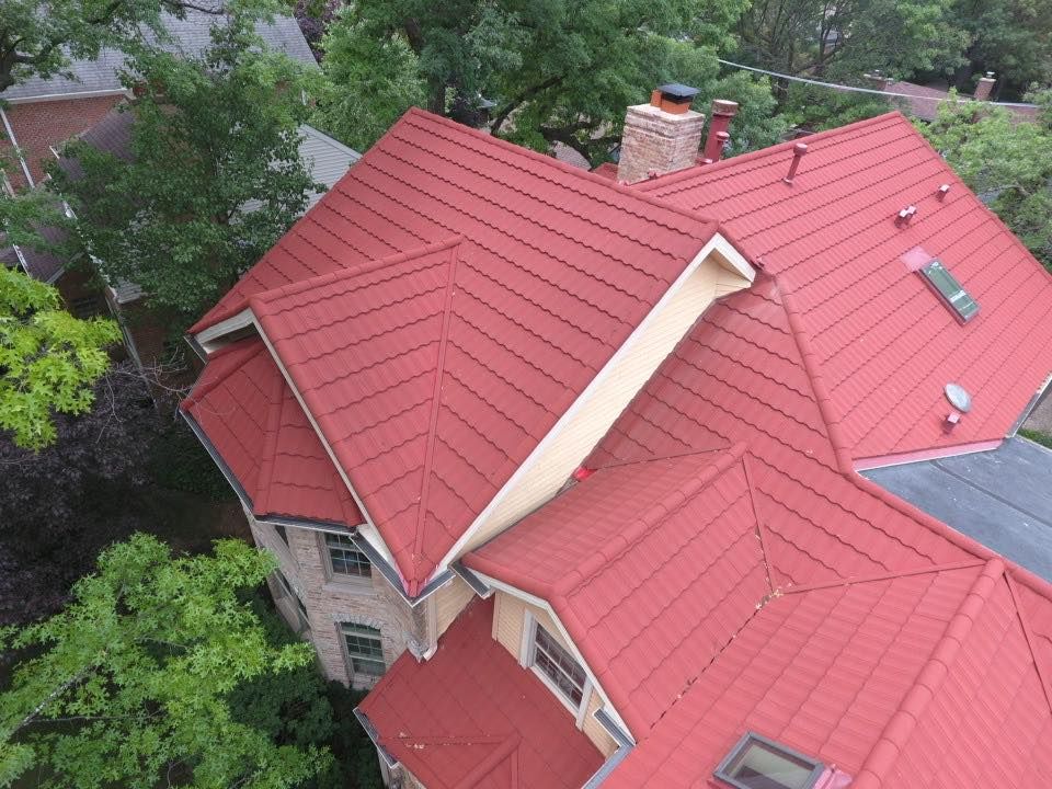 Red metal roof on a house, viewed from above, surrounded by green trees.