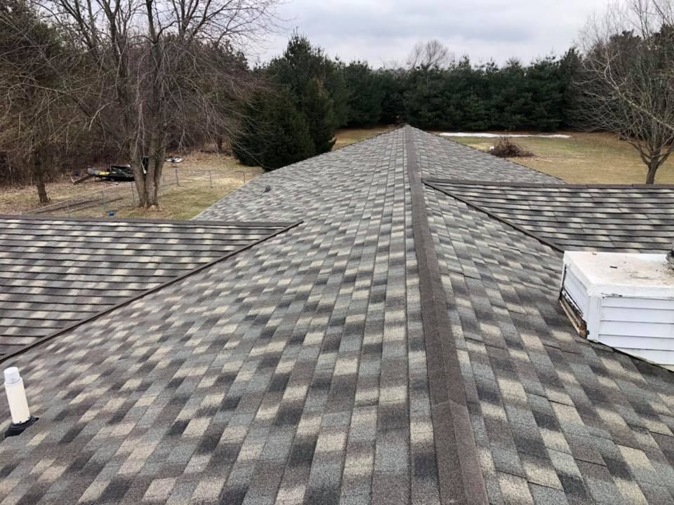 Overhead view of asphalt shingle roof, various grey and black shades, surrounded by trees and lawn.