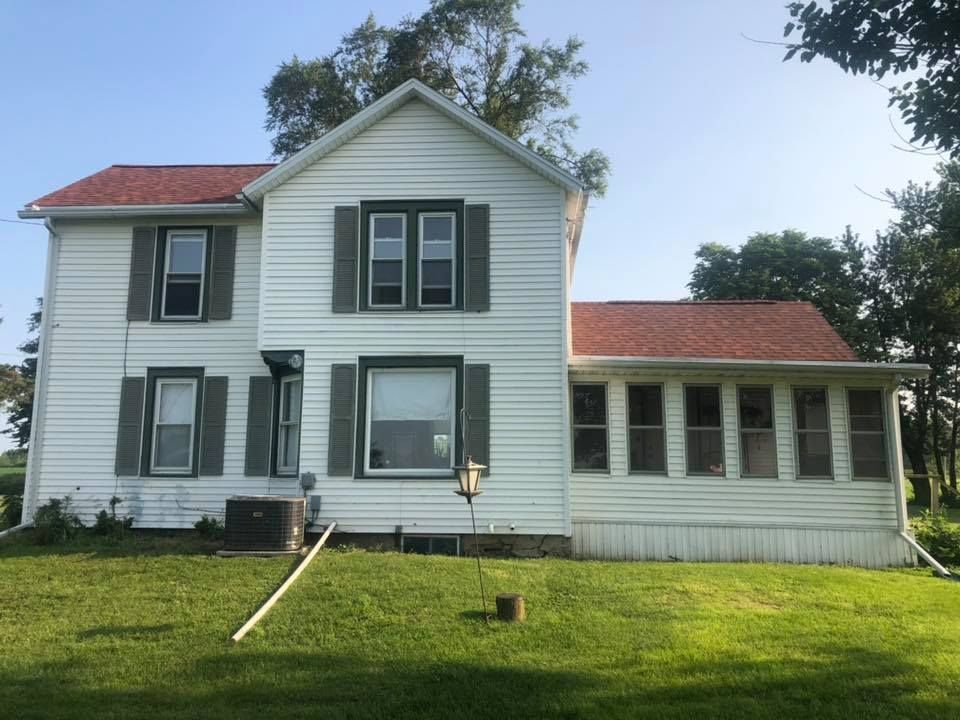 White farmhouse with red roof, green shutters, and a sunroom on a grassy lawn.