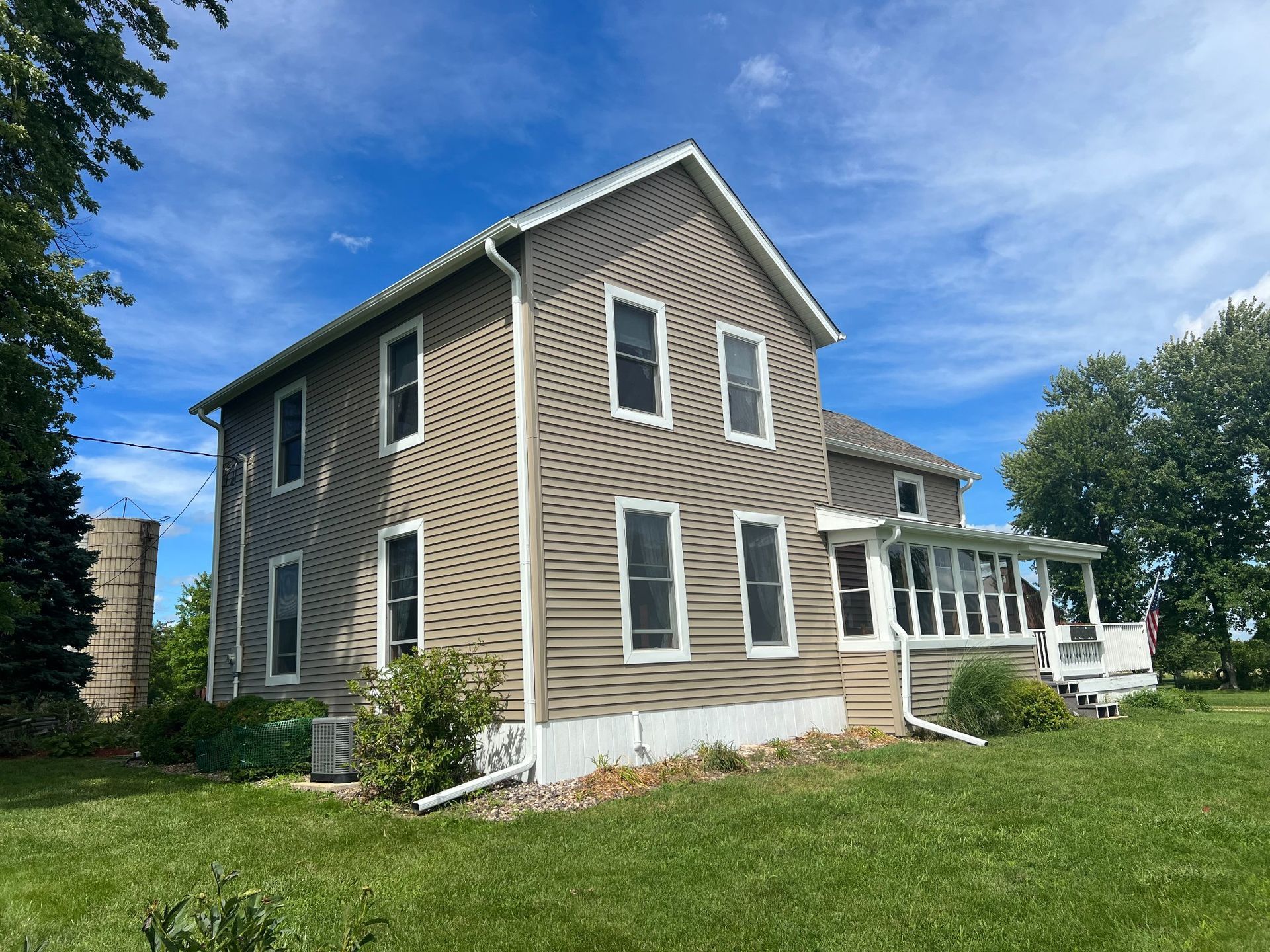 Two-story house with tan siding, white window trim, and a screened porch on a grassy lot under a blue sky.