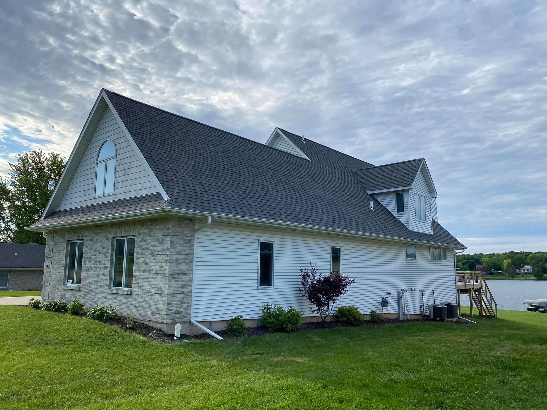 Two-story house with gray shingles, white siding, and brick accents on a green lawn near a lake under a cloudy sky.
