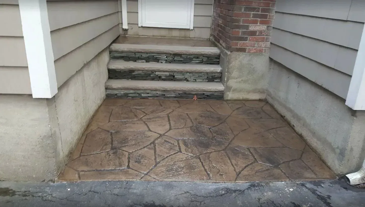 Stone-patterned concrete patio and steps leading to a doorway, set under an eave with a brick column and siding.