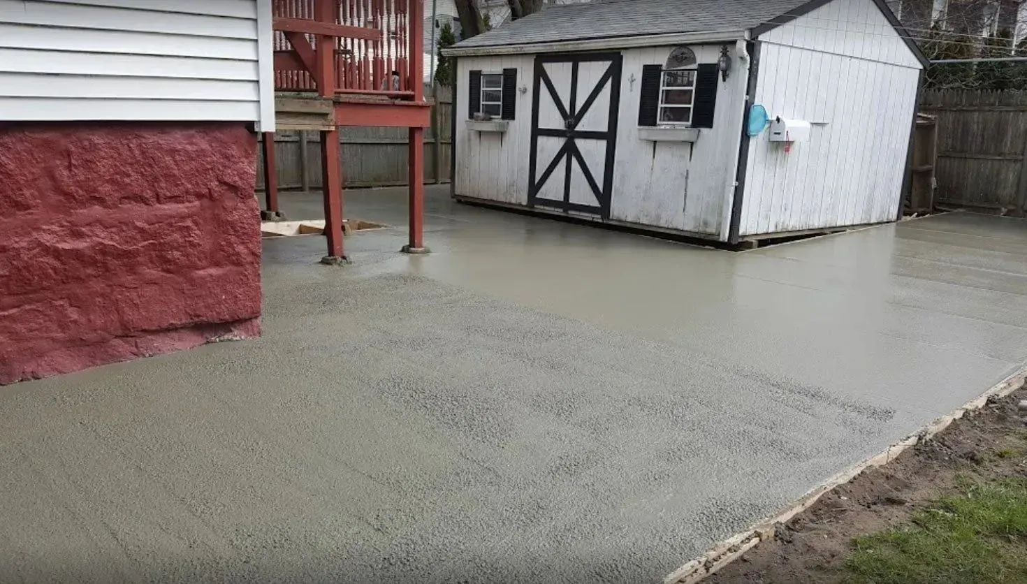 A newly poured concrete patio in a backyard setting, next to a shed and red brick wall.