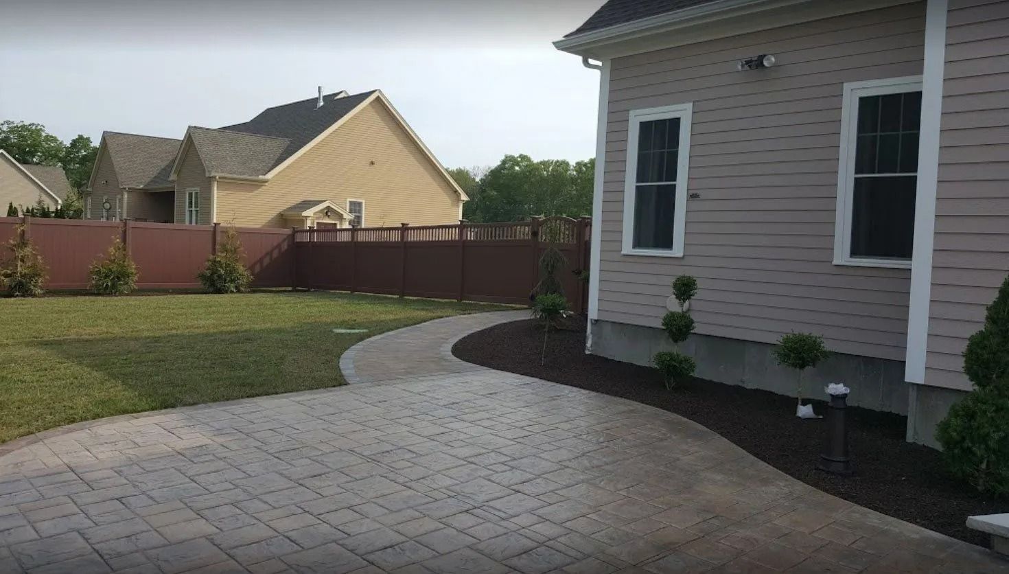 A paved path leads to a house with tan siding. A green lawn and brown fence are also visible.