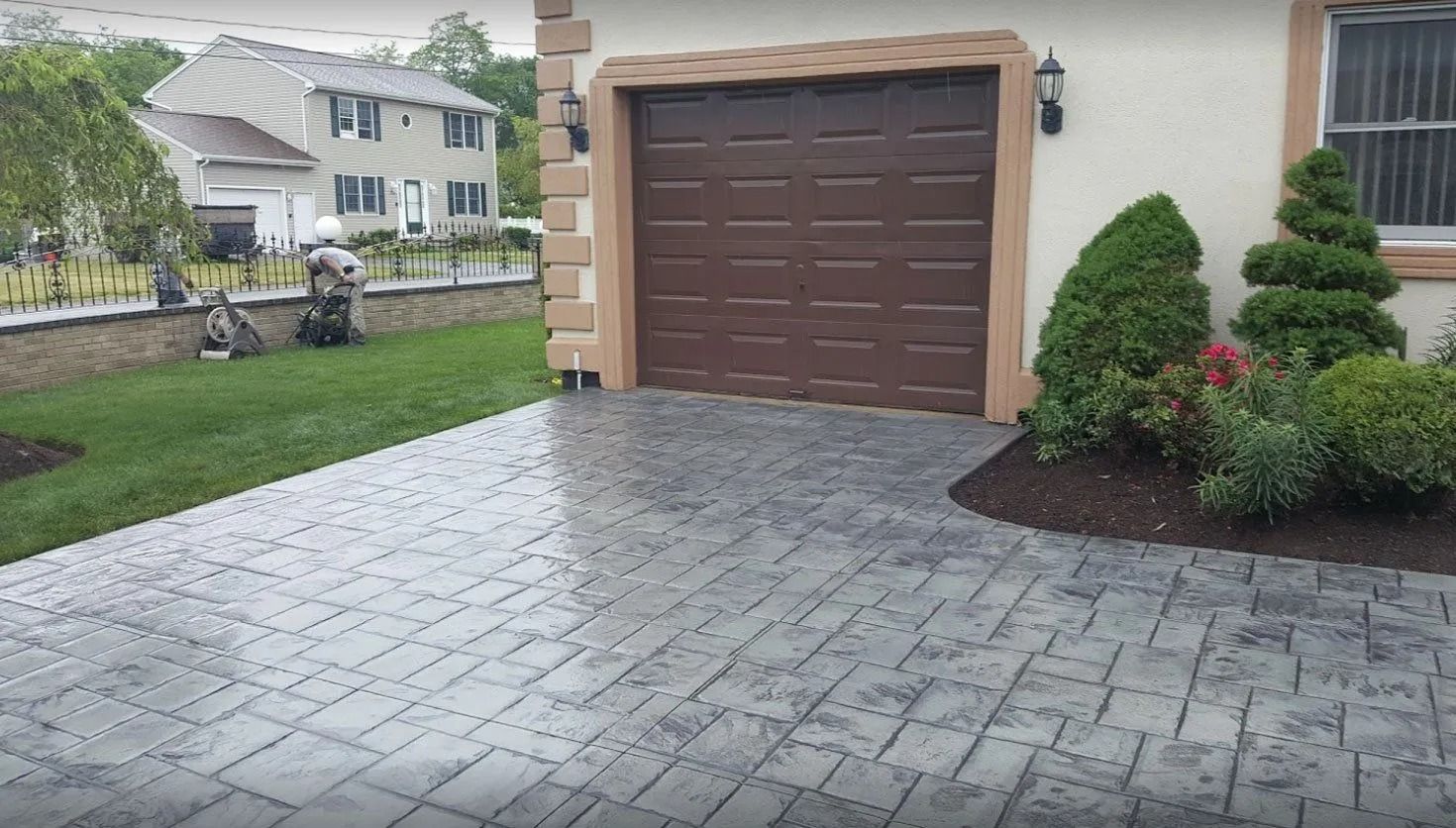 Gray patterned driveway leads to a brown garage door on a tan house with manicured landscaping.