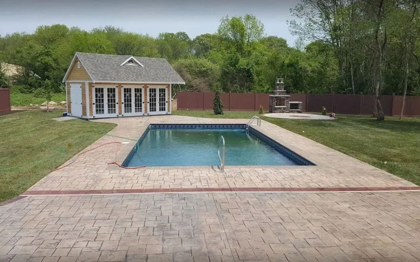 Backyard with a rectangular pool, shed, outdoor fireplace, and stamped concrete patio surrounded by trees.