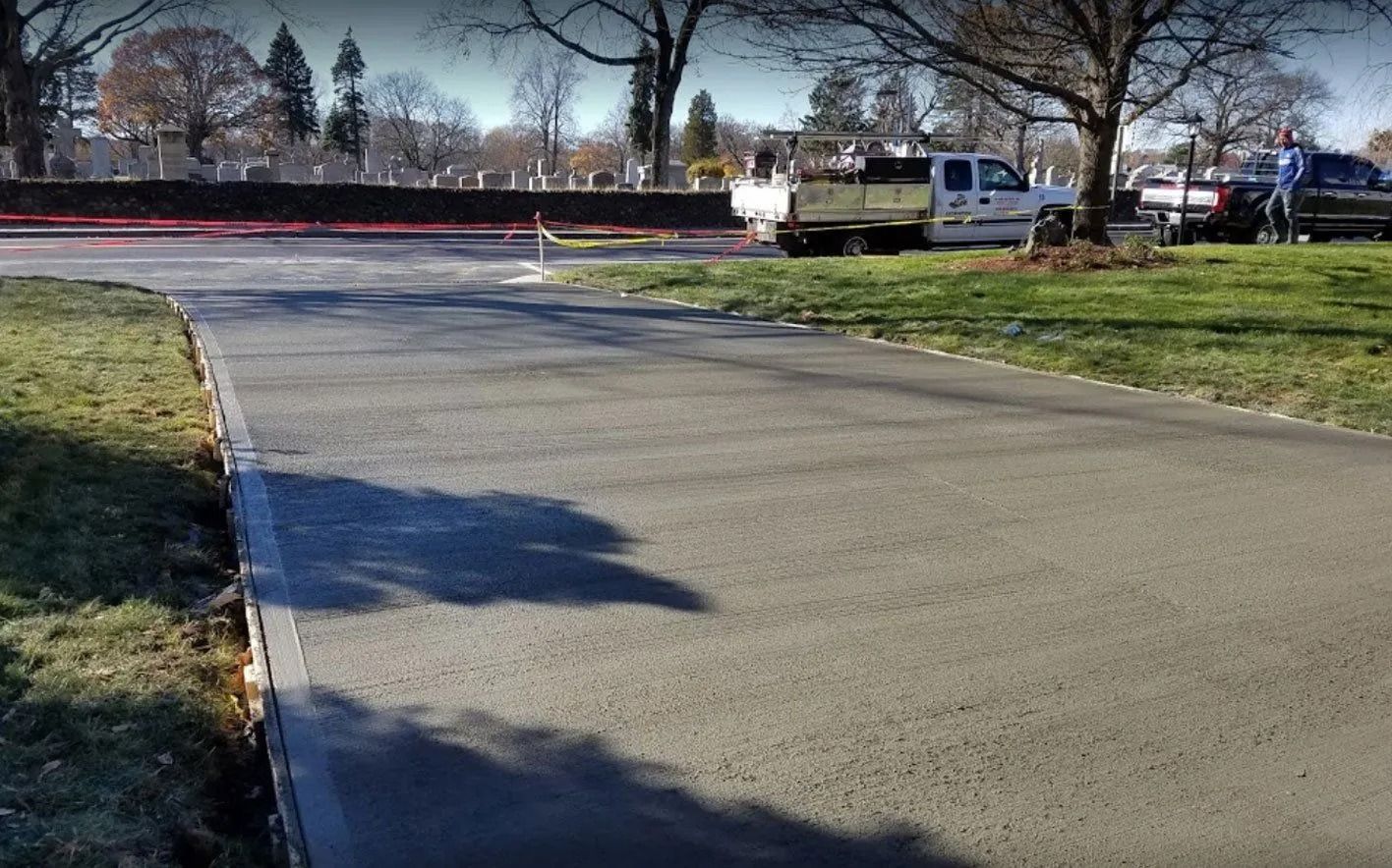 Asphalt road curves through a grassy area with vehicles parked near trees; cemetery in the background.