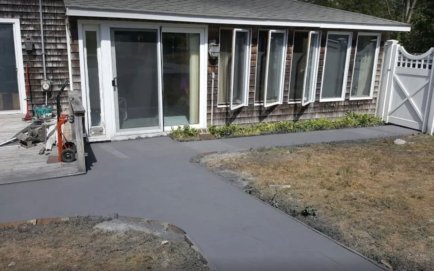 Gray concrete path leading to a house with windows. Dry grass on the right and a sliding glass door.