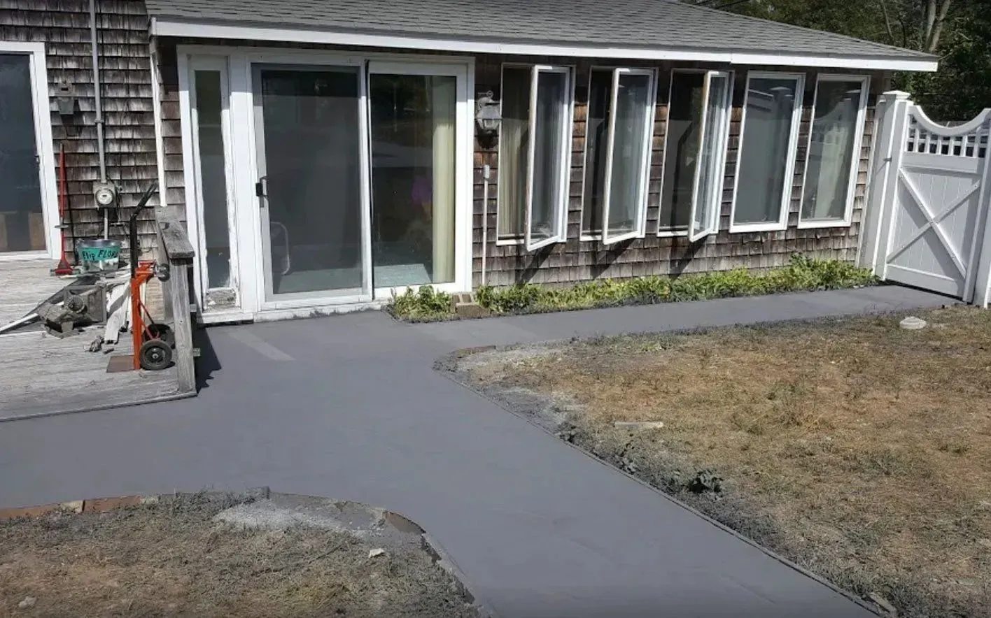 Newly poured gray concrete pathway leads to a house with a sliding glass door and multiple windows.
