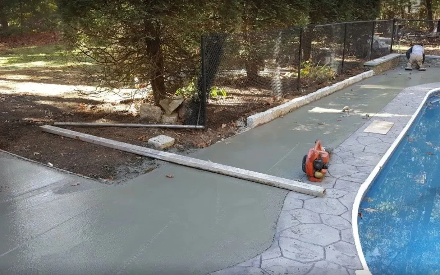 Concrete walkway being poured around a pool; worker using a blower.