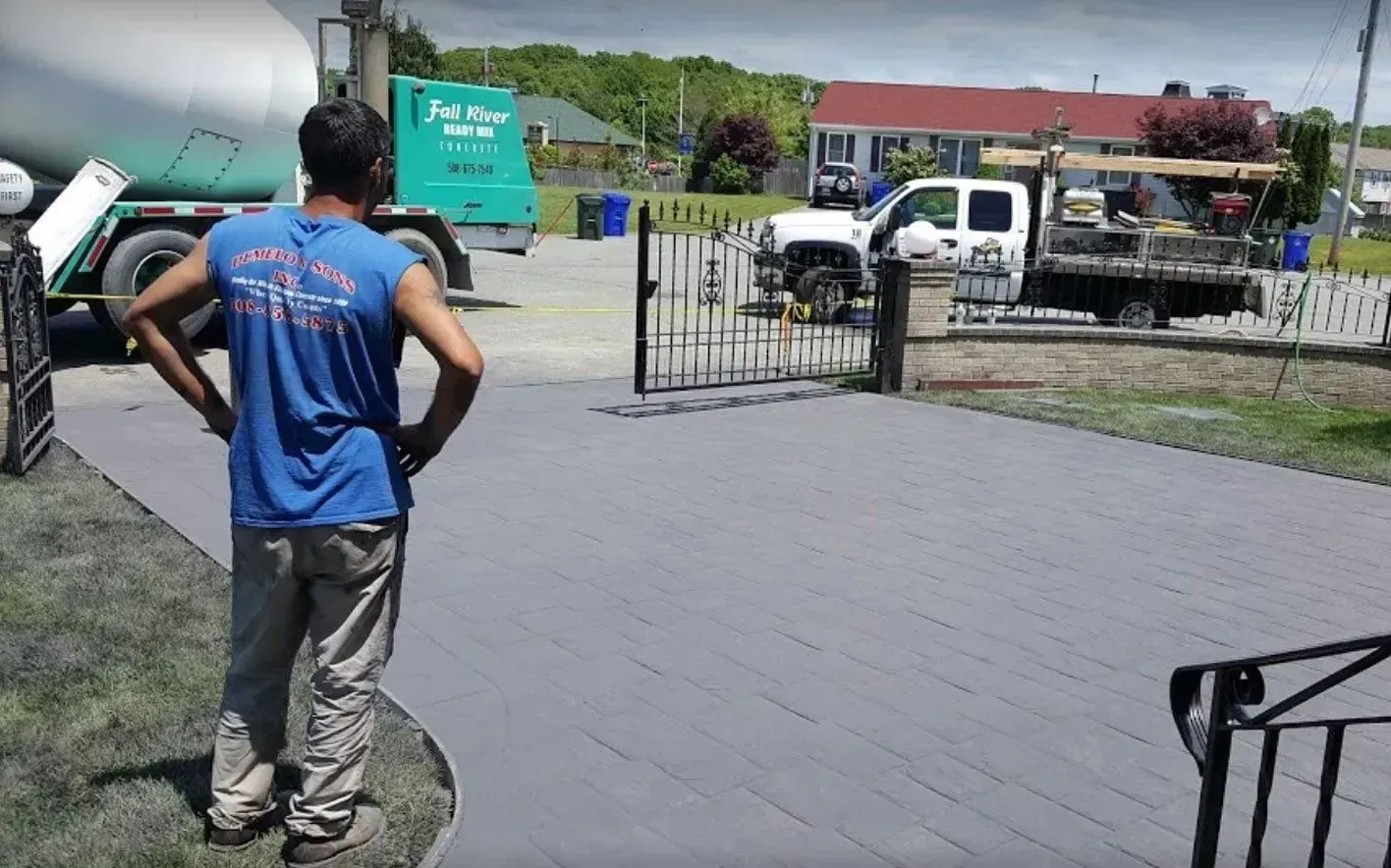 Man standing, hands on hips, surveys a newly paved gray driveway; work vehicles visible.