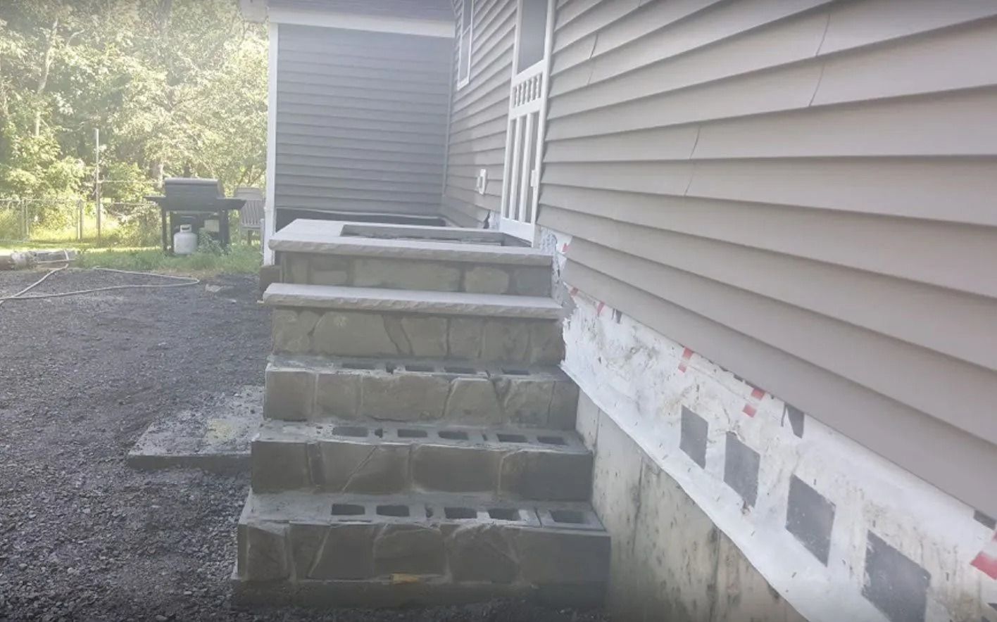 Stone steps leading up to a house with gray siding. Unfinished construction visible at the base.
