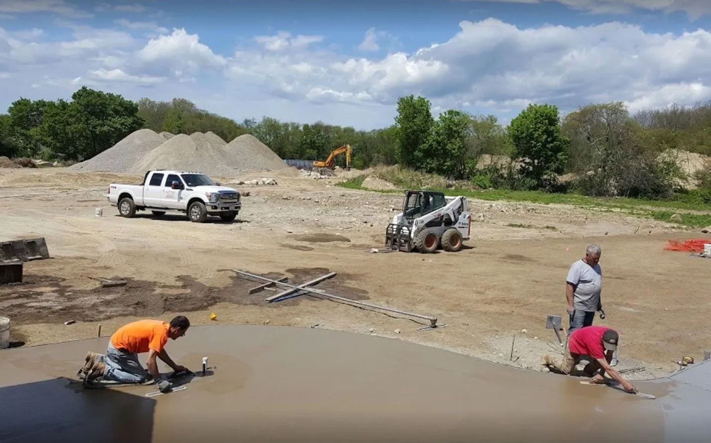 Construction workers leveling wet concrete; a truck, skid steer, and excavator are in the background.