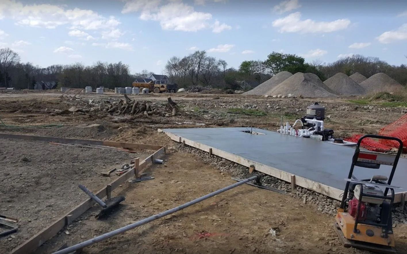 Construction site with freshly poured concrete slab. Equipment and piles of dirt visible in background.