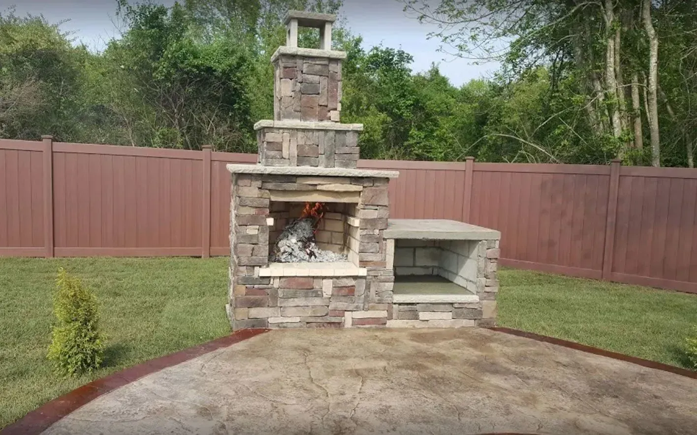 Stone outdoor fireplace and cooking area in a backyard. Fire burning inside. Brown fence and green grass in background.