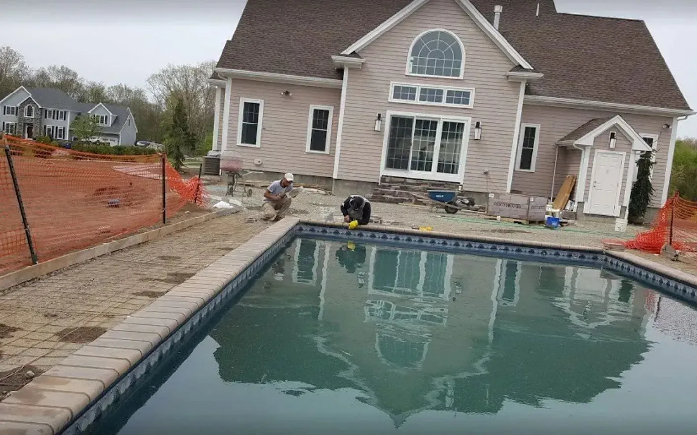 House with pool under construction; workers near pool edge. Building is light-colored.
