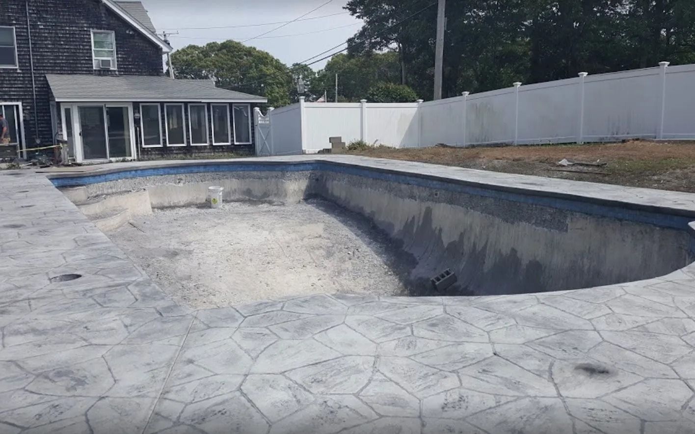 An empty concrete swimming pool in a backyard, surrounded by a house, fence, and trees on a sunny day.