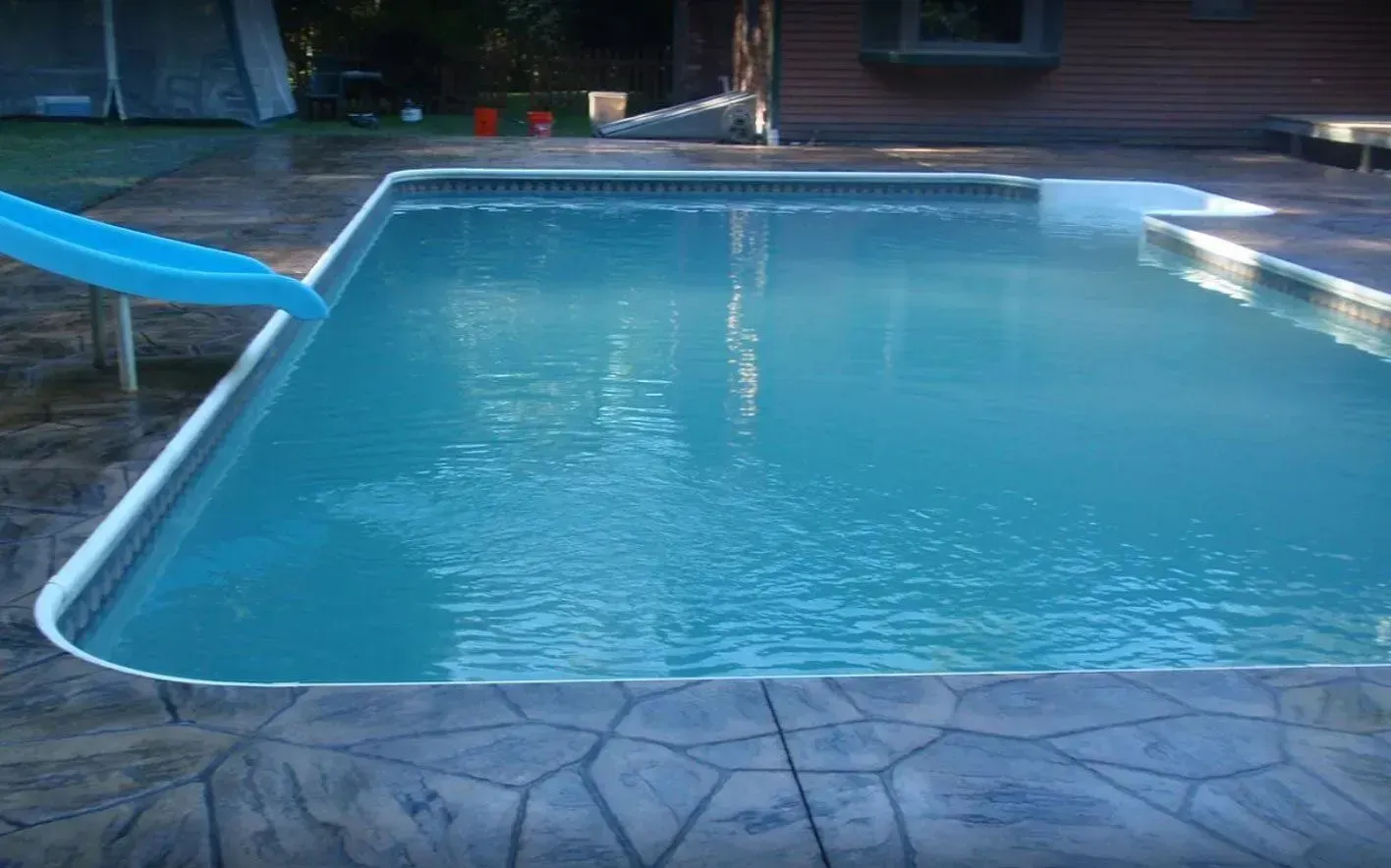 Blue swimming pool with a slide, surrounded by a stone-like patio.