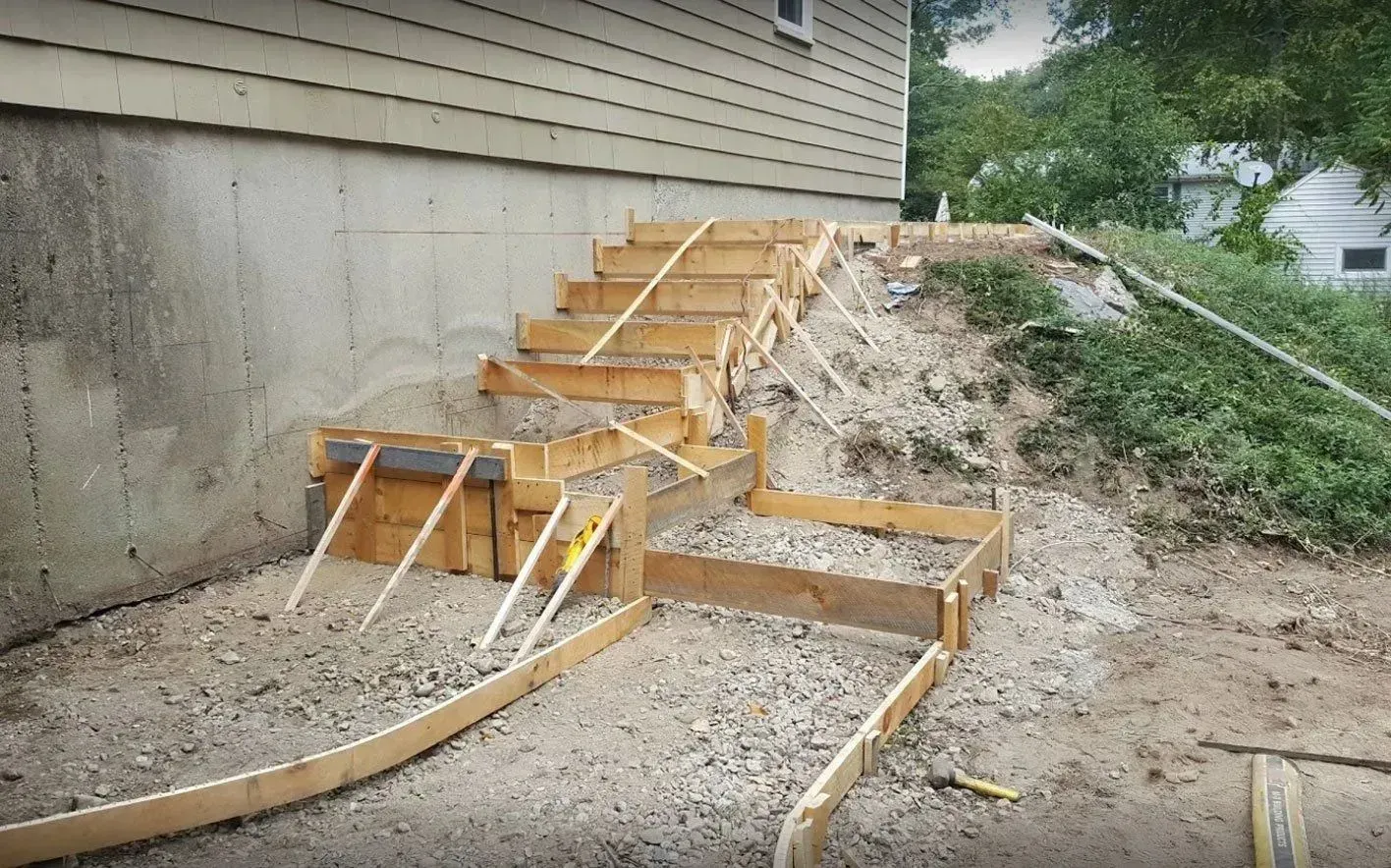 Wooden forms for concrete stairs being built beside a concrete foundation and sloped ground.