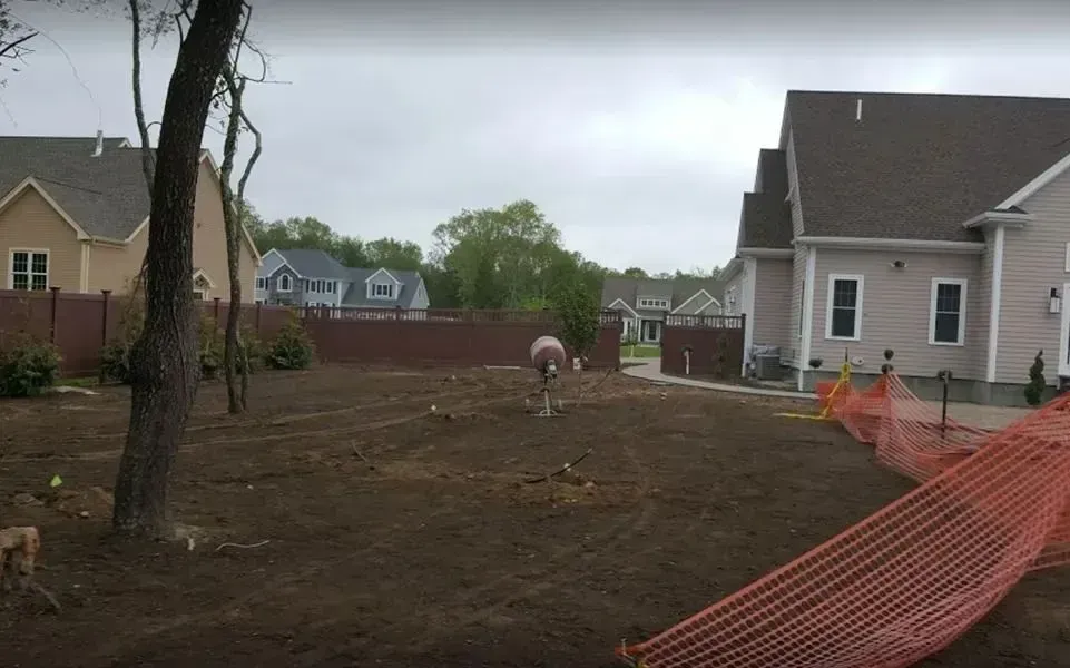 Brown backyard with a newly tilled garden bed and fencing. Houses are in the background, orange construction fencing in the foreground.