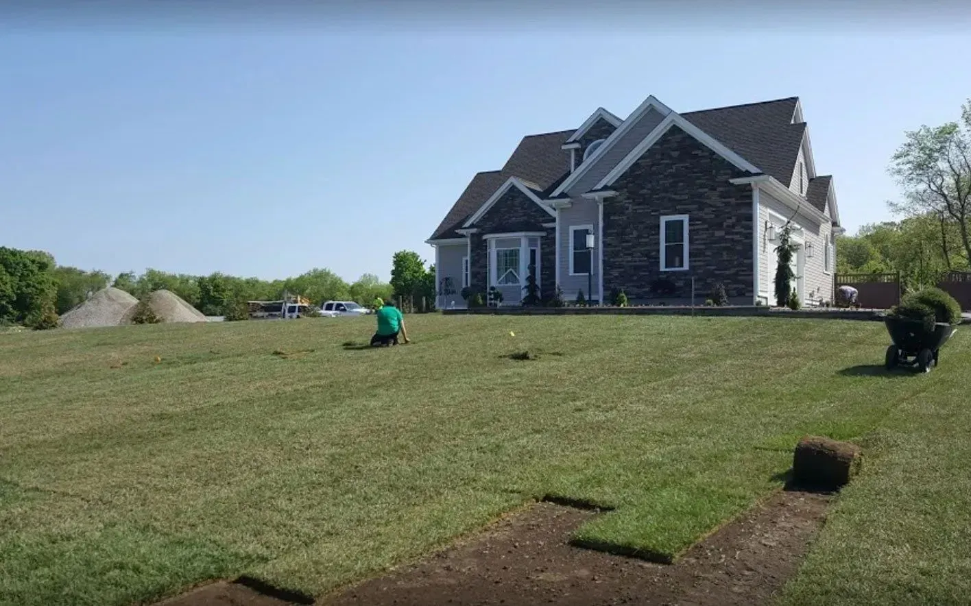 Laying sod in front of a house. Person placing sections of green grass, blue sky, piles of dirt in the background.