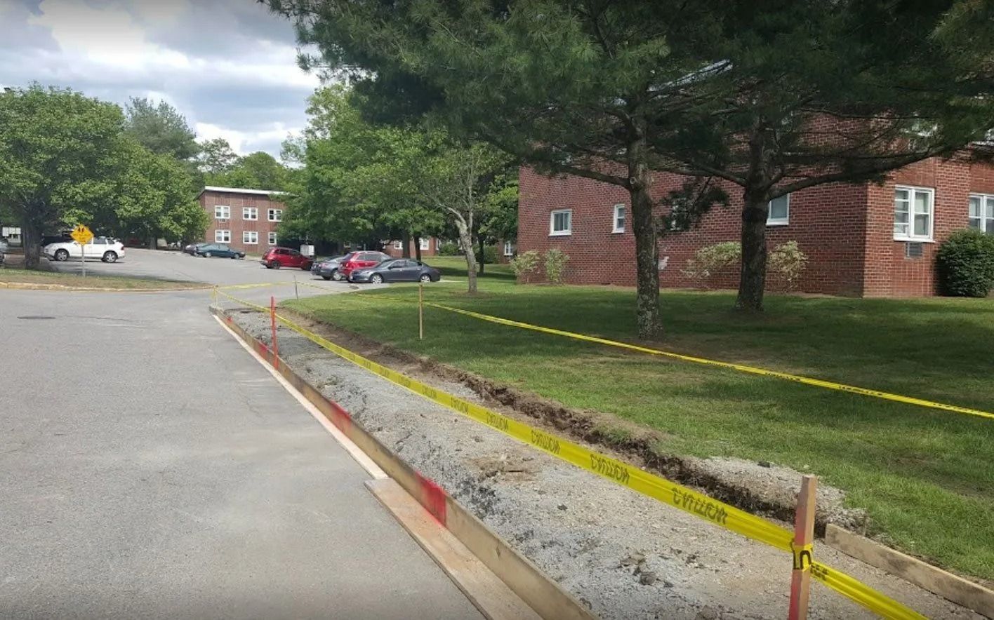 Roadside construction with a trench, warning tape, and parked cars. Buildings and trees in the background.