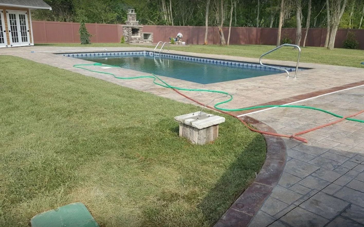 Backyard with pool, lush green grass, patio, and trees. Hoses are laid out.