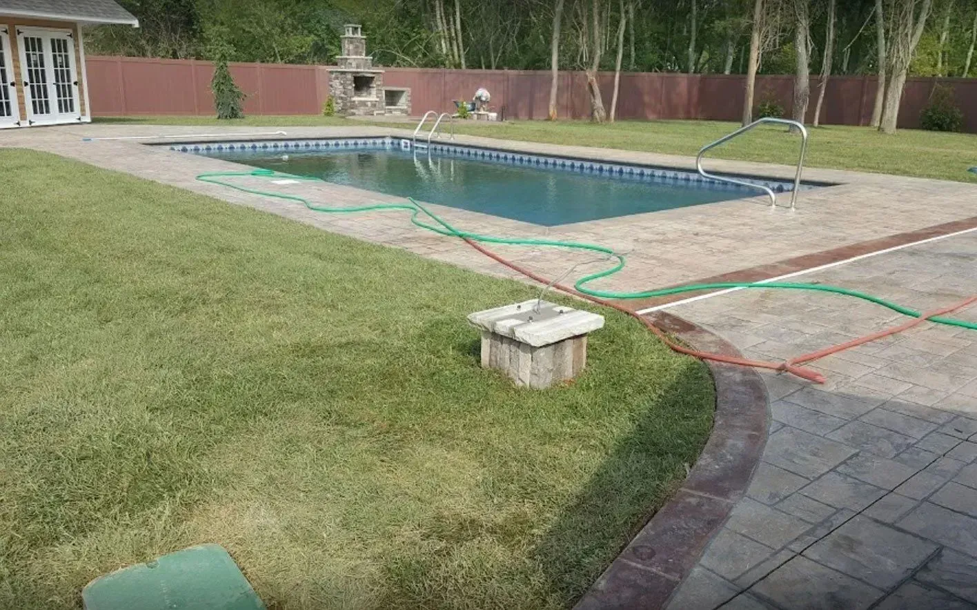 A rectangular swimming pool surrounded by a concrete patio and lawn, with hoses and a small well-like structure nearby.