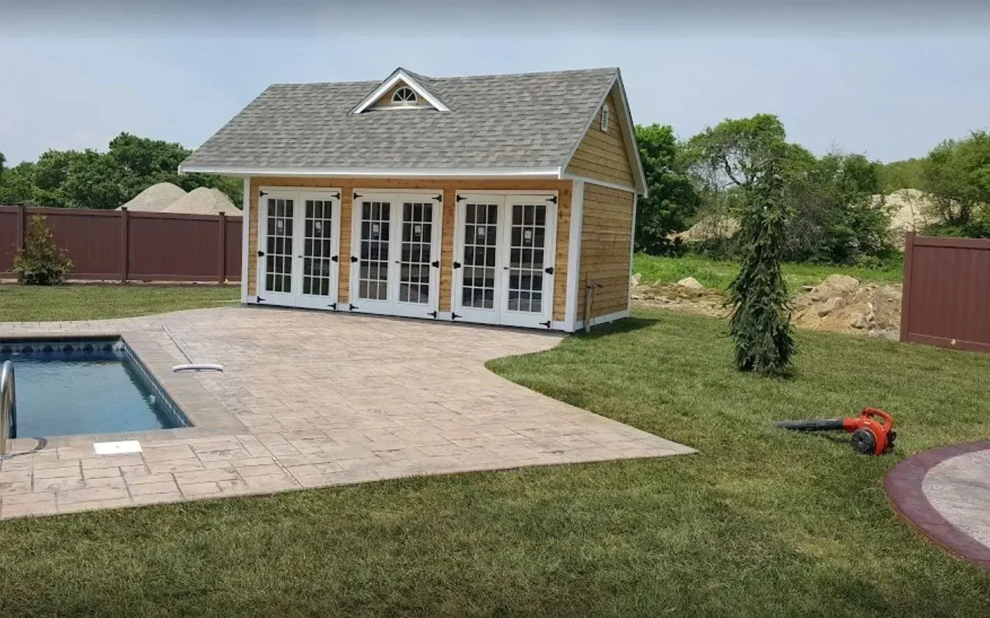 Poolside shed with French doors, gray roof, brown fence, and stamped concrete patio.