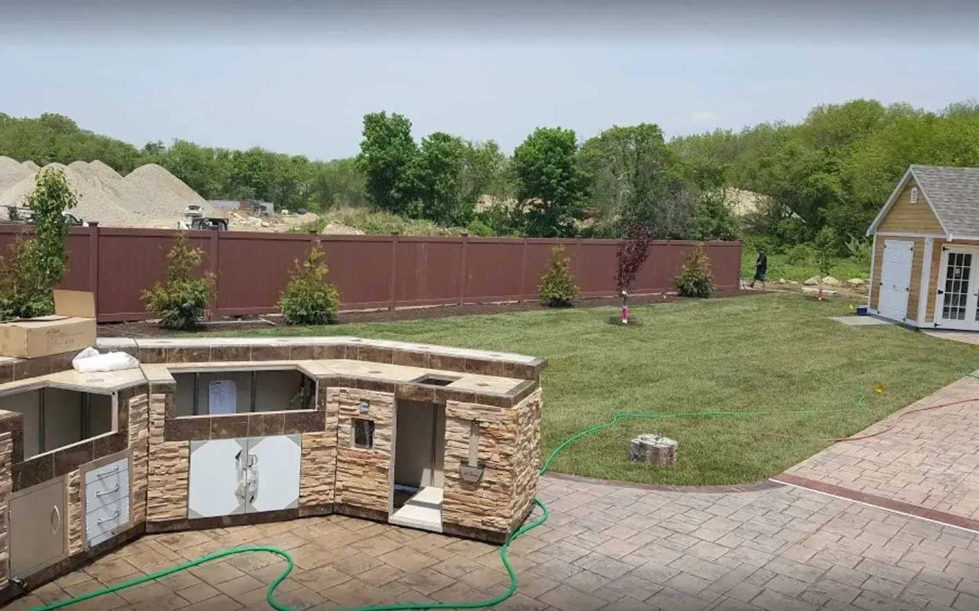 Outdoor kitchen with brown fence, small trees, green lawn, and a small yellow shed.