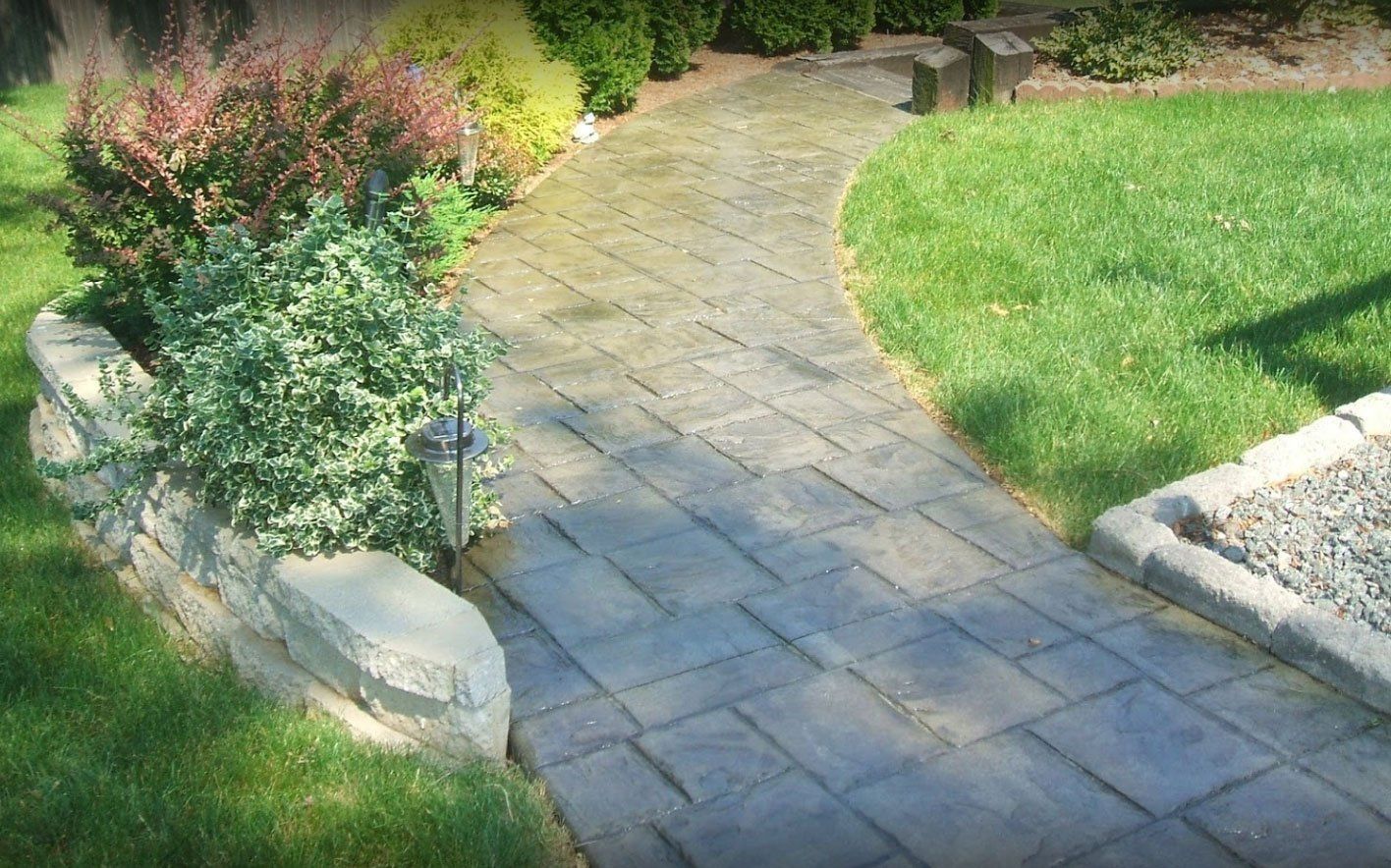 Brick pathway winding through a green lawn, edged by a stone wall with bushes.