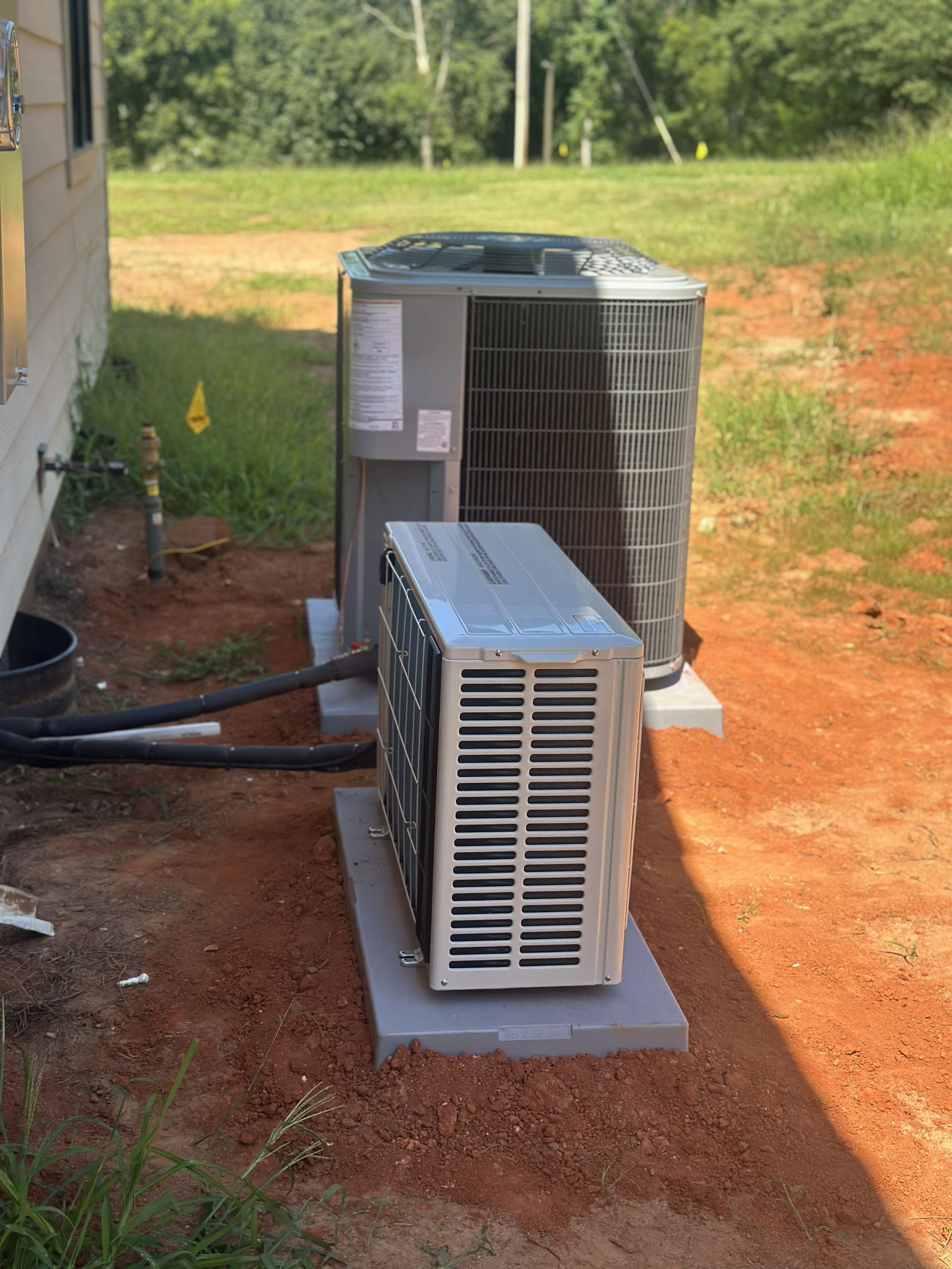 Two HVAC outdoor units sit on concrete pads on red dirt next to the side of a house.