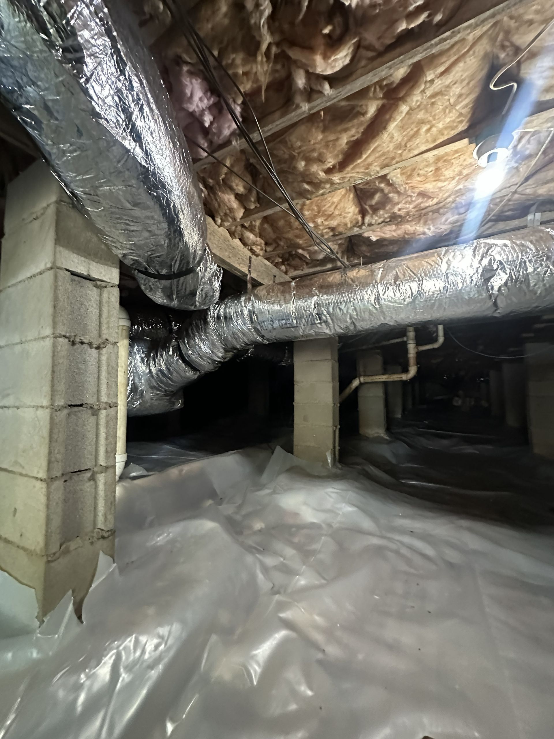 A crawl space with a plastic vapor barrier on the ground, support pillars, and insulated HVAC ductwork overhead.