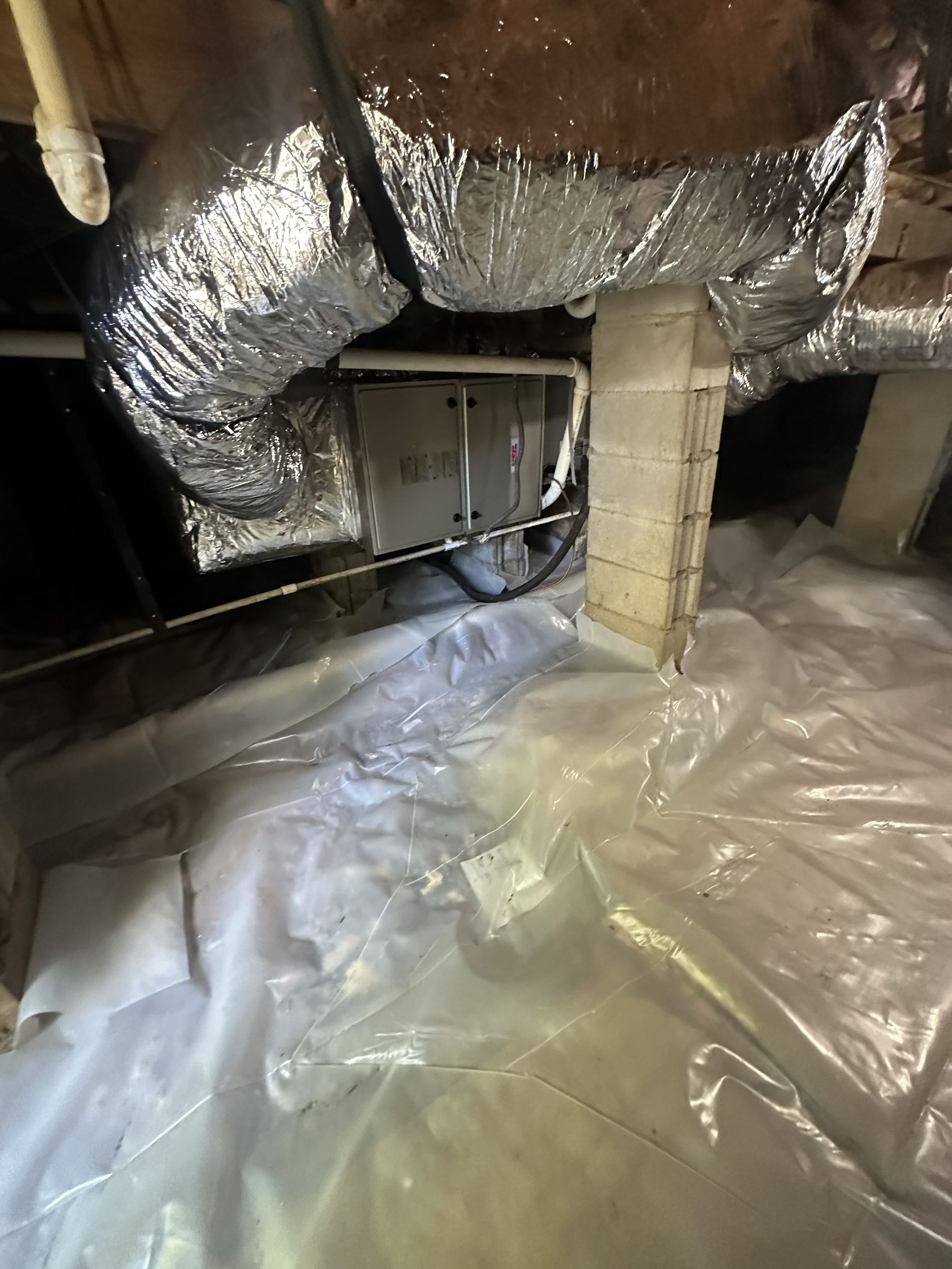 A crawl space featuring a metal HVAC unit and foil-wrapped ducts above a white plastic vapor barrier on the ground.