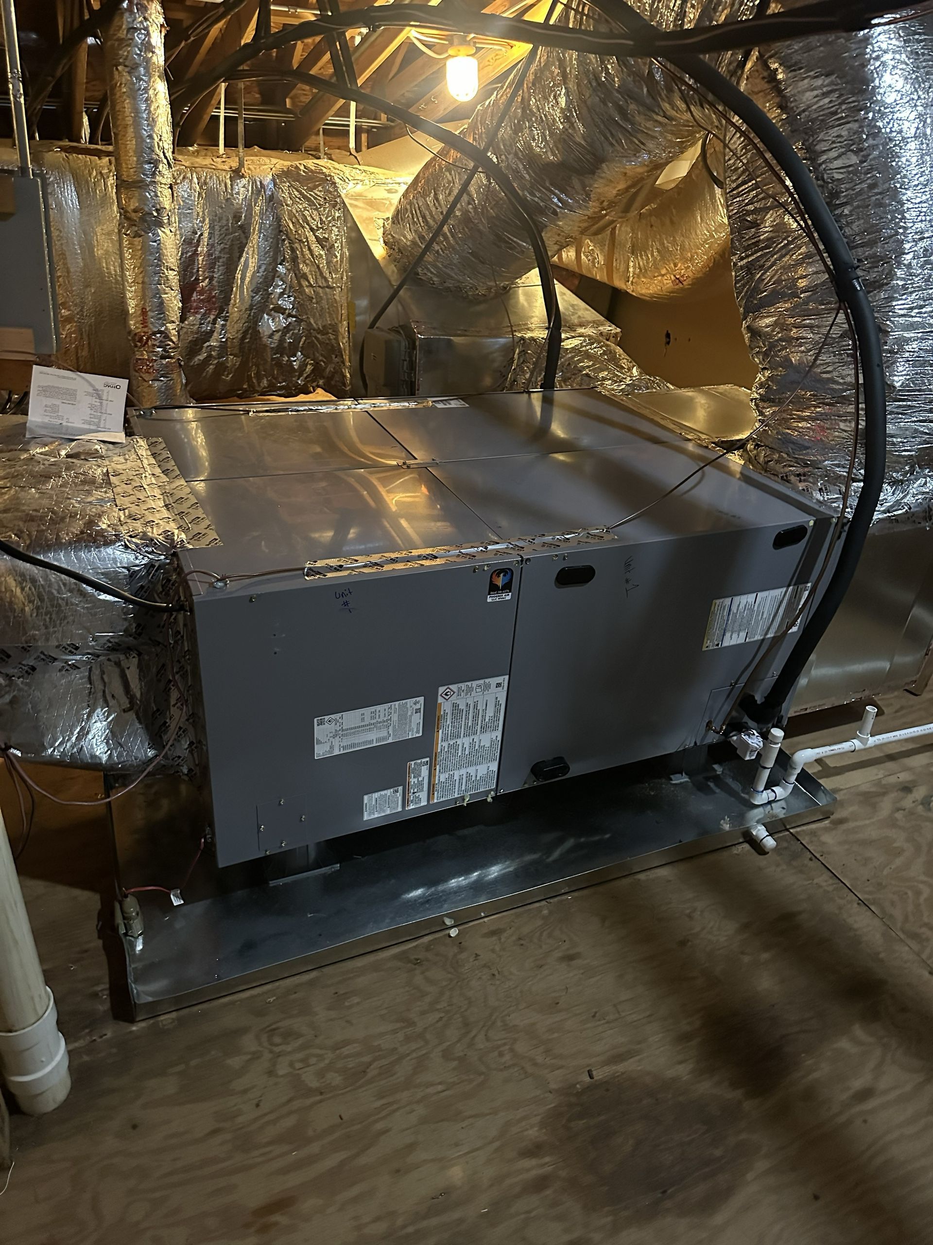 An HVAC air handler unit installed in an attic, with silver metallic ductwork and a condensate drain pan.